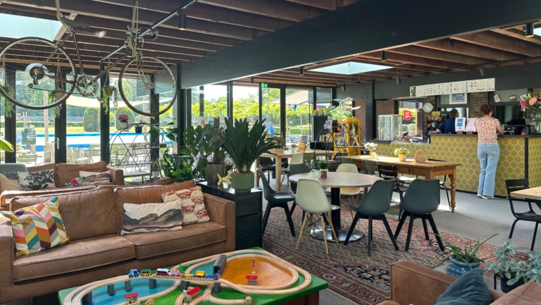 Interieur van een café met houten plafond en veel ramen. Er zijn bruine leren banken, een houten tafel met stoelen, en een speelgoedtrein op een tafeltje. Aan het plafond hangen fietsen en er zijn veel planten en decoratieve elementen. Een persoon staat bij de toonbank.