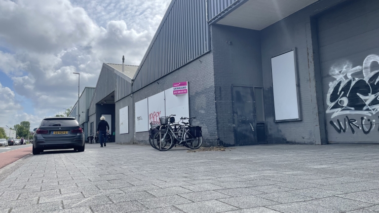 Industrieel gebouw met donkere bakstenen, fietsen geparkeerd tegen de muur, auto geparkeerd aan de linkerkant, en graffiti op de muur. Een bord met "VERHUURD" is zichtbaar boven de fietsen. De lucht is bewolkt.