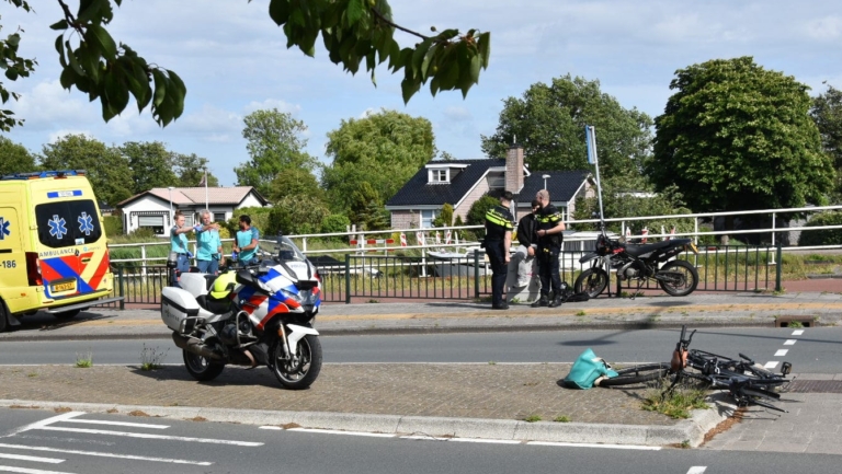Een verkeersongeval met een politieagent op een motorfiets, een ambulance, en een beschadigde fiets op de grond. Ambulancepersoneel en politieagenten zijn aanwezig bij het incident dat zich afspeelt op een weg met een brug en omringd door groen.