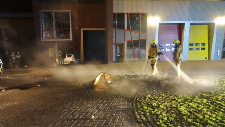 Twee brandweermannen in beschermende kleding blussen een brand op straat, met rookwolken rond hen, voor een gebouw met rode en gele deuren 's nachts.