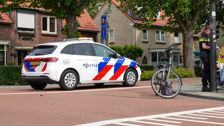 Een politiewagen staat geparkeerd op een straat, met een politieagent die naast een fiets op de stoep staat. Op de achtergrond zijn huizen met rode dakpannen en bomen te zien.