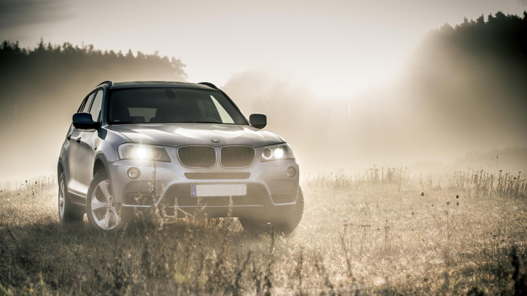 Een zilveren SUV met koplampen aan, rijdend over een grasveld in een mistige omgeving met bomen op de achtergrond.