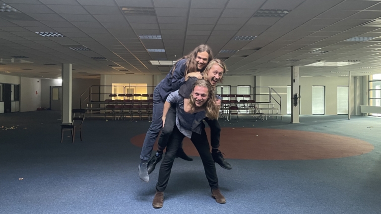 Drie personen staan in een lege zaal, met twee van hen bovenop elkaar gestapeld als een menselijke piramide.