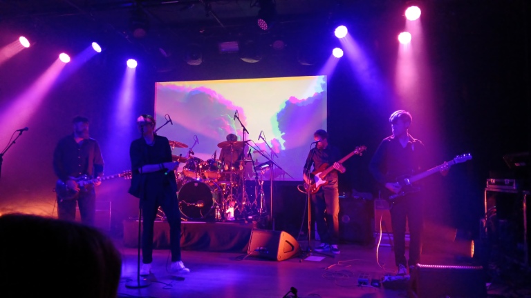 Een band treedt op op een podium met felgekleurde verlichting en een scherm met abstracte beelden op de achtergrond. Drie gitaristen, een drummer en een zanger zijn te zien, gehuld in donkergekleurde kleding.