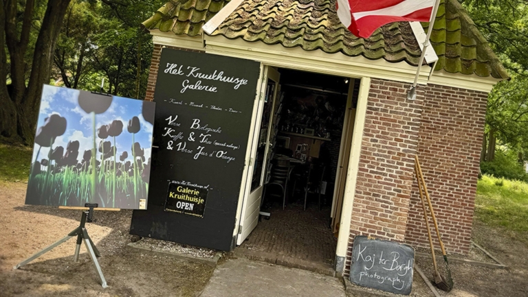 Een kleine galerie genaamd 'Het Kruithuisje' met een bord dat koffie, thee en vers sapjes aanbiedt. Voor de galerie staat een schilderij van bloemen en hangt een rood-witte vlag aan de gevel.