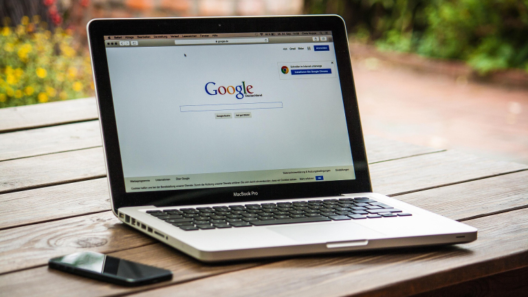 Een MacBook Pro op een houten tafel met de Google startpagina op het scherm, naast een smartphone.