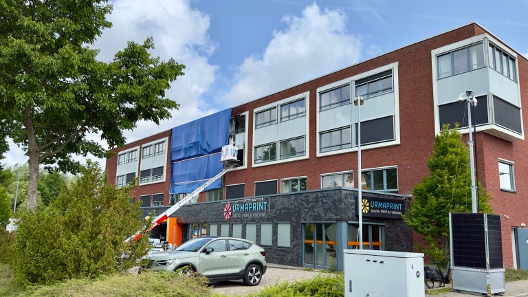 Een bakstenen bedrijfsgebouw met meerdere verdiepingen, waarbij een deel bedekt is met een blauw zeil. Er staat een hoogwerker met een persoon bezig aan het gebouw. Op de begane grond is er een bedrijf genaamd 'Urmaprint'. Voor het gebouw staat een geparkeerde auto en er zijn bomen en struiken zichtbaar.