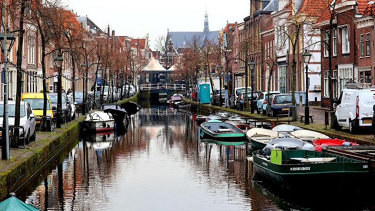 Een kanaal in Nederland met aan weerszijden geparkeerde auto's en oude huizen, en verschillende boten aangemeerd langs de kade.