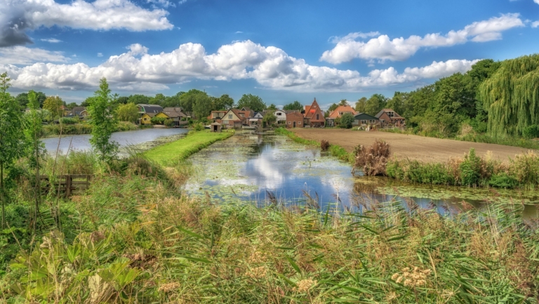 Landschap in Nederland met grachten, weelderig groen en op de achtergrond traditionele huizen onder een blauwe lucht met wolken.