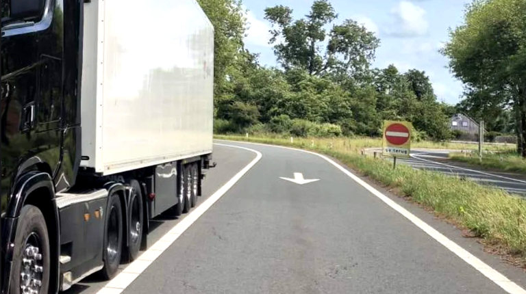 Een zwarte vrachtwagen rijdt op een landweg omgeven door bomen en groen; een verkeersbord met een verbodsteken en de tekst "ga terug" staat langs de weg.