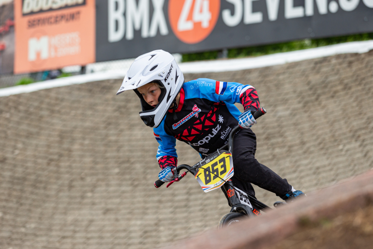 Een kind met een witte helm en blauw-rode BMX-outfit rijdt op een BMX-fiets met nummerbord 853 op een baanhelling, gefocust op de weg voor zich.