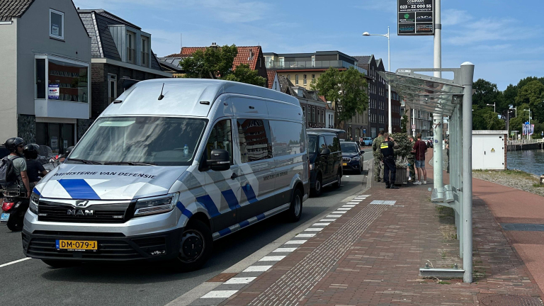 Een grijs busje van het Ministerie van Defensie geparkeerd langs de weg in een stedelijke omgeving, met een politieagent en een paar mensen ernaast bij een bushalte langs een kanaal.