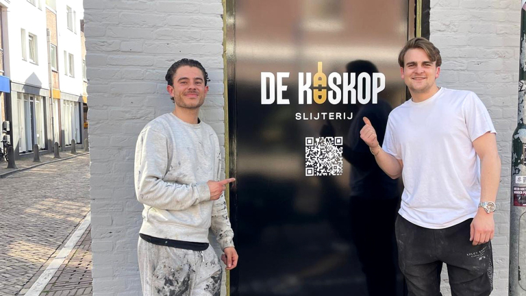 Twee mannen in vrijetijdskleding wijzen naar een gevel met het logo en de naam 'De Kaskop Slijterij' en een QR-code, in een stedelijke straatomgeving.