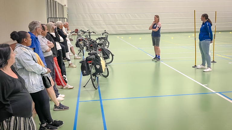 Een groep mensen in een sporthal kijkt naar twee instructeurs die uitleg geven; fietsen staan in een rij naast hen.