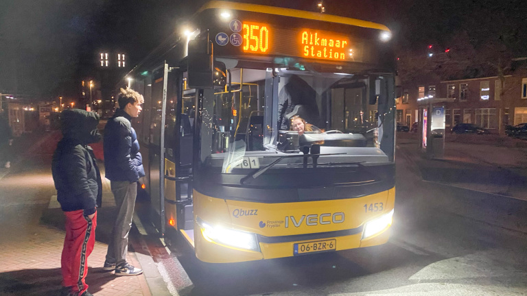 Twee mensen staan op een donkere avond bij een geparkeerde gele bus met de bestemming Alkmaar Station, met het nummer 350 op de voorruit. De bus is van het merk Iveco en heeft het logo van Qbuzz en Provincie Fryslân.