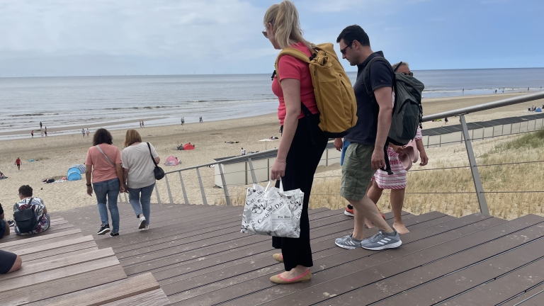 Mensen lopen op een houten trap naar het strand, met de zee op de achtergrond. Enkelen dragen rugzakken en tassen. Op het strand zijn parasols en mensen zichtbaar. Het weer lijkt bewolkt.