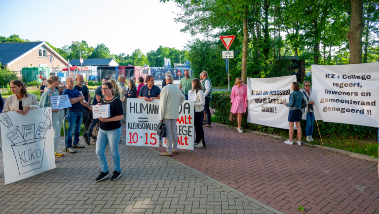 Groep mensen houdt protestborden vast tijdens een demonstratie op een stoep met bomen en gebouwen op de achtergrond.