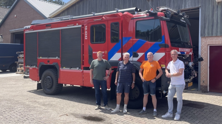 Vier mannen staan buiten naast een brandweerwagen bij een gebouw met een houten gevel.