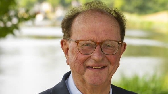 Ouderling met bril en pak, glimlachend buiten bij een rivier.