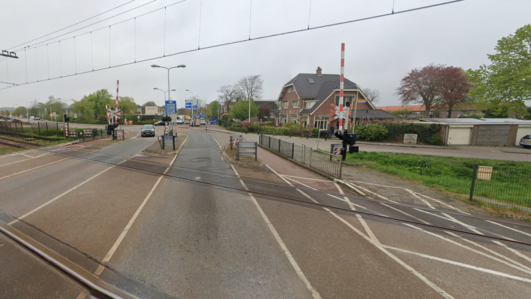 Een spoorwegovergang met slagbomen die omhoog staan, een auto die het spoor nadert, en een straat met woningen aan de rechterkant. Fietspaden en verkeersborden zijn zichtbaar op de achtergrond, samen met bomen en een grijze lucht.