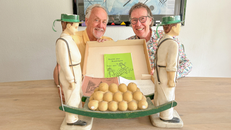 Twee lachende mensen poseren achter een houten beeld van kaasdragers met een doos gevuld met historische kaasdragersboekjes en papieren op de voorgrond.
