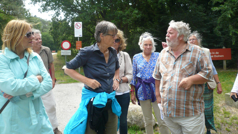 Een groep mensen staat buiten in een bosrijke omgeving, in gesprek. Sommige personen dragen kleurrijke kleding en zonnebrillen. Er zijn verkeersborden en een informatiebord op de achtergrond.