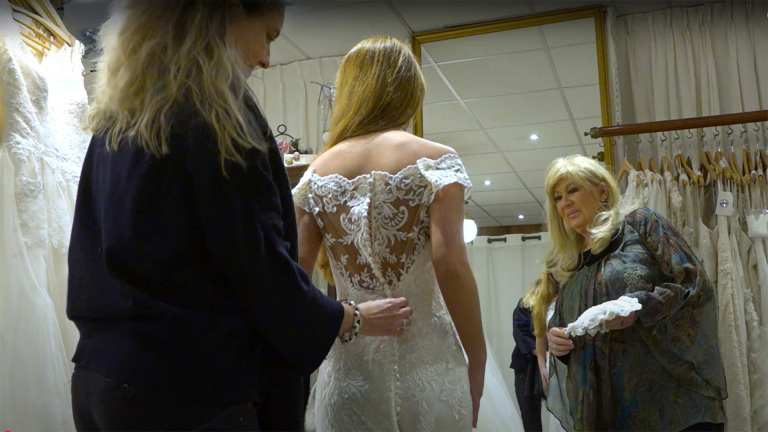 Een vrouw in een bruidswinkel past een kanten trouwjurk met open rug, terwijl twee andere vrouwen haar helpen en toekijken.