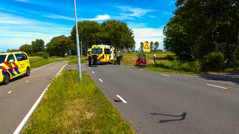 Ambulance en noodvoertuig op een weg bij een verkeersongeluk met een rode scooter aan de kant, omringd door paramedici en een verkeersbord met een waarschuwing voor een kruispunt in de achtergrond.