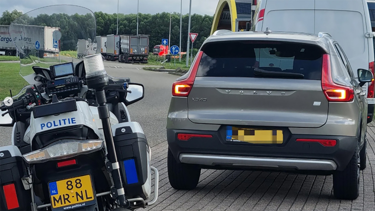 Een politie-motorfiets naast een geparkeerde zilverkleurige Volvo XC40 op een parkeerplaats met vrachtwagens op de achtergrond.