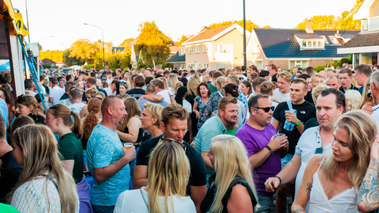 Een grote menigte mensen op straat tijdens een evenement genaamd Tour de Waard “Marlene”, op een zonnige dag met huizen op de achtergrond.