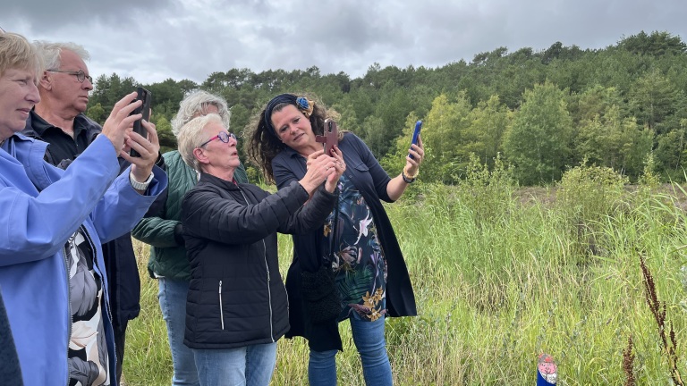 Een groep mensen staat in een veld met gras en bomen op de achtergrond, terwijl ze met hun telefoons foto's maken of iets bekijken.