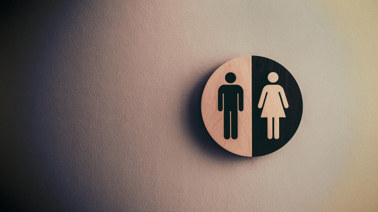 Een rond houten bord met daarop de symbolen voor mannen en vrouwen, gesplitst in twee helften, tegen een muur bevestigd.