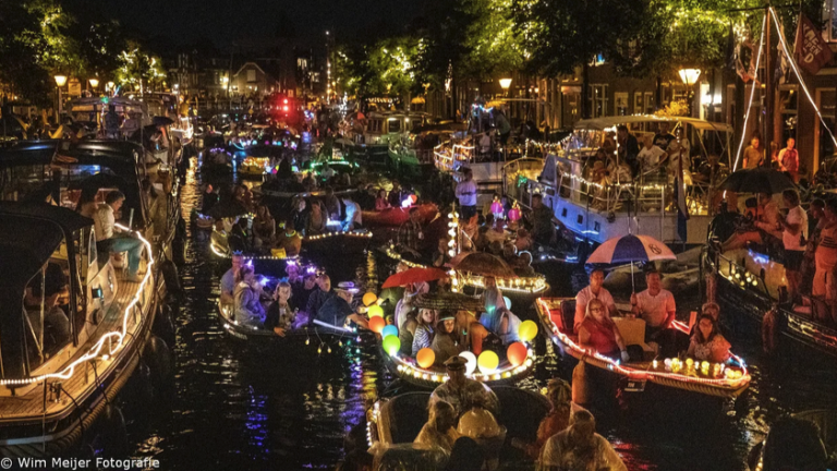 Een druk bezochte gracht in de avond, vol met kleurrijk verlichte boten en mensen onder paraplu's, omringd door sfeervolle straatverlichting en silhouetten van bomen.
