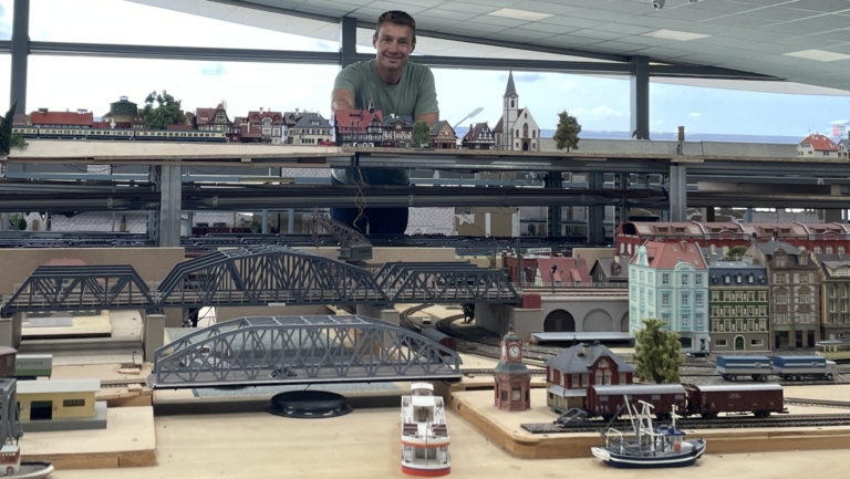 Een persoon staat achter een uitgebreide modelspoortafel met gedetailleerde miniatuurgebouwen, bruggen en treinen in een overdekte ruimte met glazen wanden.