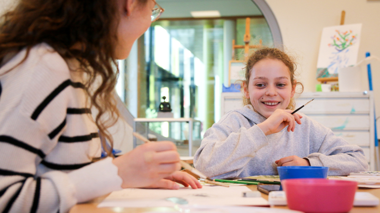 Twee meisjes tekenen en schilderen aan een tafel in een lichte ruimte, lachend en bezig met schildermaterialen en papier.