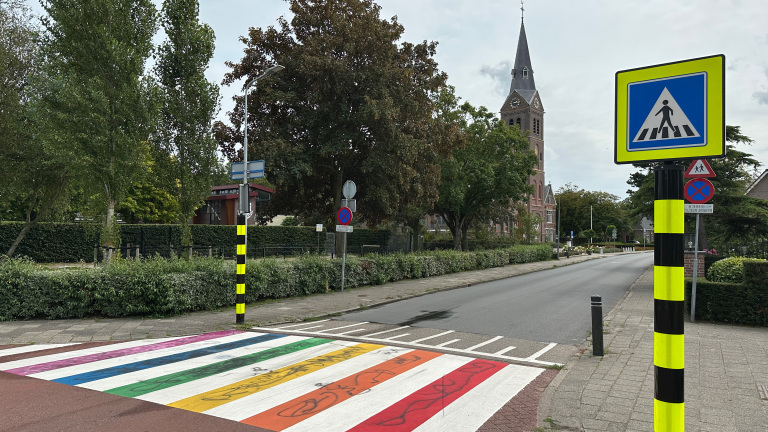 Straatbeeld met een regenboogzebrapad en verkeersborden in een groene omgeving, met een kerk op de achtergrond.