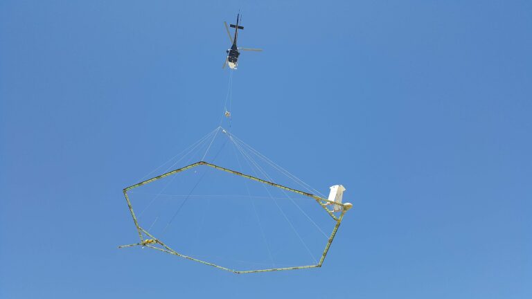 Een helikopter vervoert een groot, zeshoekig geofysisch meetinstrument in de lucht tegen een heldere blauwe hemel.