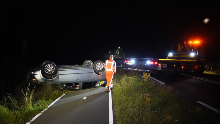 Een gekantelde auto ligt op zijn dak op een weg 's nachts, terwijl een persoon in oranje veiligheidskleding naar de scene loopt. In de achtergrond staat een takelwagen met geopende lichten.