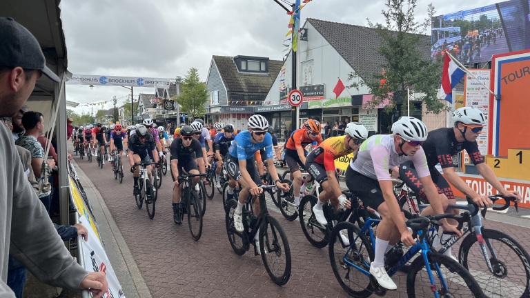 Groep wielrenners tijdens een wedstrijd in een stadsomgeving, omringd door toeschouwers en reclameborden.