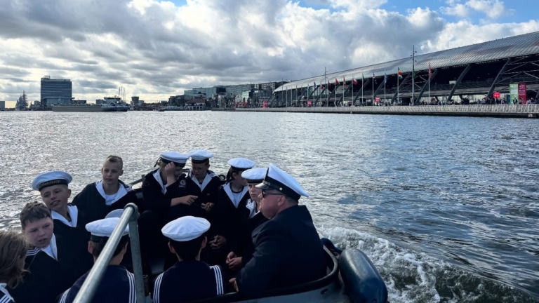 Kleine groep zeekadetten in marine-uniformen met witte petten zitten in een boot op het water, met een grote overdekte terminal rechts en gebouwen aan de horizon onder een bewolkte lucht.