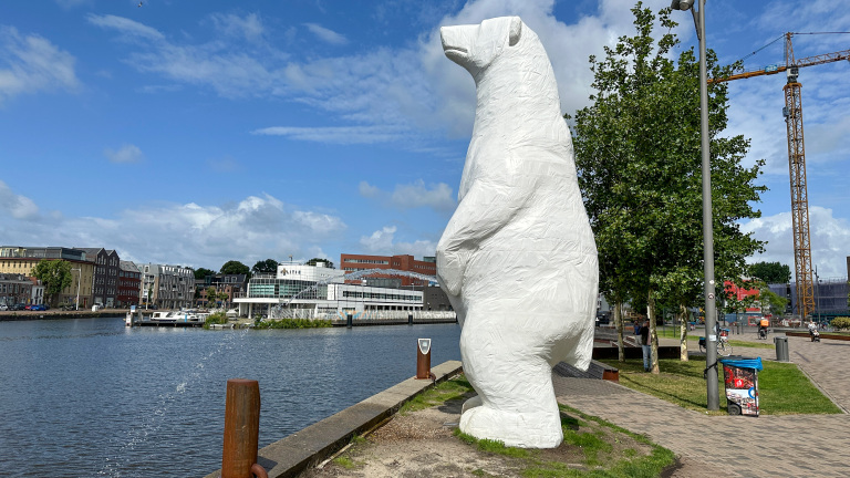 Een grote, witte ijsbeerstandbeeld van tape staat langs een kanaal met gebouwen, bomen en een hijskraan op de achtergrond onder een blauwe lucht met wolken.