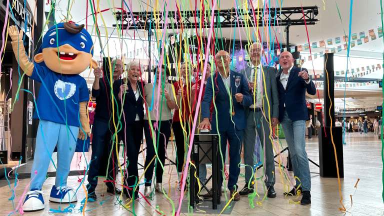 Een groep mensen, waaronder een mascotte in een blauw kostuum, staan te vieren met confetti in een winkelcentrum.