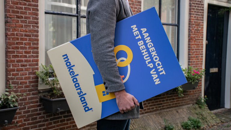 Een persoon in een grijze jas draagt een blauw bord met de tekst 'Aangekocht met behulp van makelaarsland,' naast een bakstenen muur en een raam.