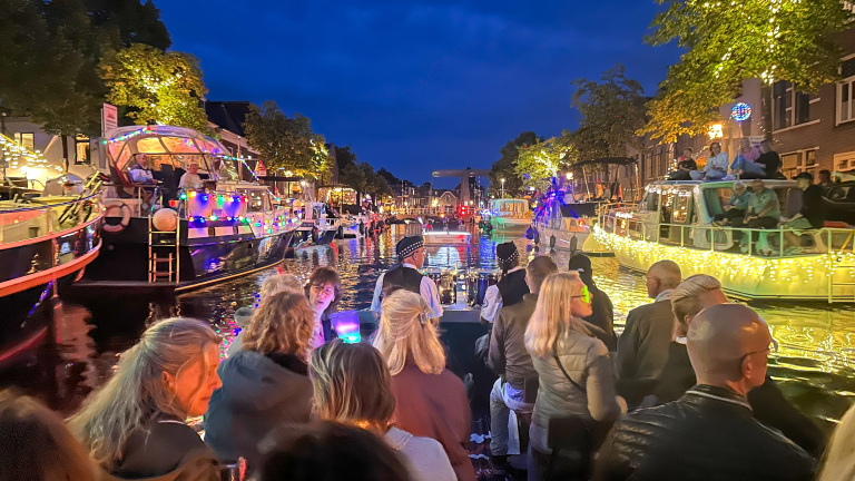 Feestelijke boten versierd met kleurrijke lichtjes varen 's avonds door een kanaal in een stad, omringd door mensen die op de boten staan en toekijken.