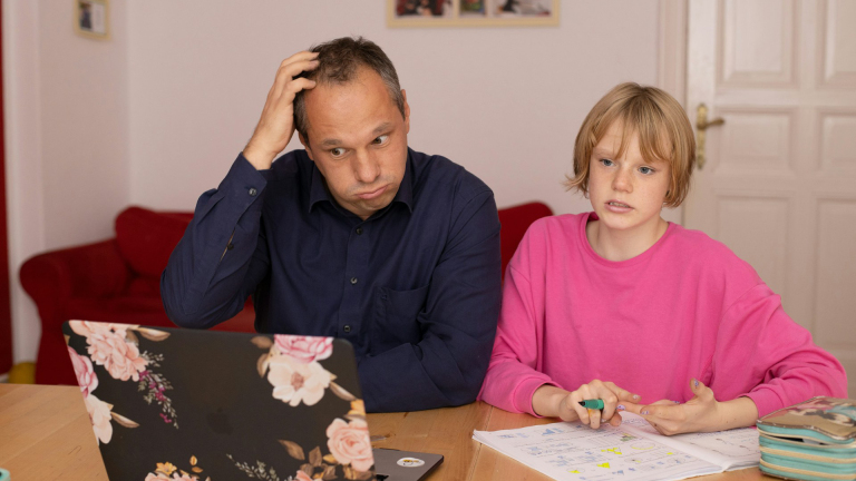Een man en een kind zitten aan een tafel met een open laptop en huiswerk voor zich; de man kijkt bezorgd terwijl het kind iets uitlegt.
