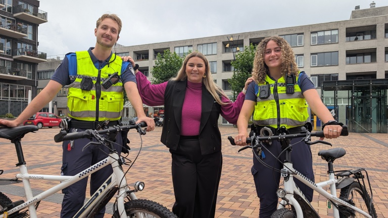 Twee handhavers in uniform staan naast een vrouw, allemaal glimlachend, met fietsen op de voorgrond en een stedelijke omgeving als achtergrond.