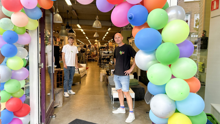 Twee mannen staan glimlachend bij de ingang van een winkel, versierd met kleurrijke ballonnen, met verschillende meubels en verlichting zichtbaar binnen.