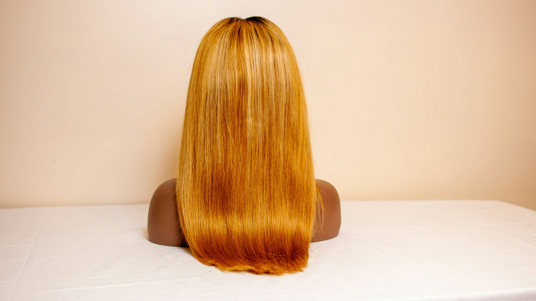 Wig met lange, rechte, gouden lokken geplaatst op een poppenhoofd, op een witte tafel tegen een neutrale achtergrond.