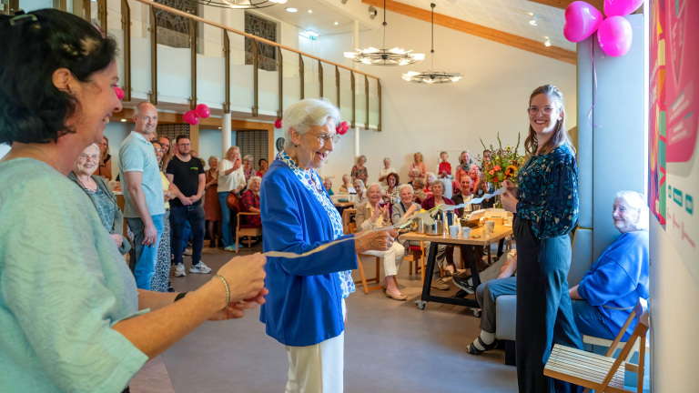Een oudere vrouw in een blauwe blazer staat glimlachend voor een groep mensen in een gemeenschapsgebouw of feestzaal.