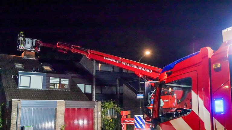 Een brandweerwagen van Brandweer Heerhugowaard staat voor een huis, met een ladder uitgeschoven naar het dak waarop een brandweerman te zien is, in de nacht.